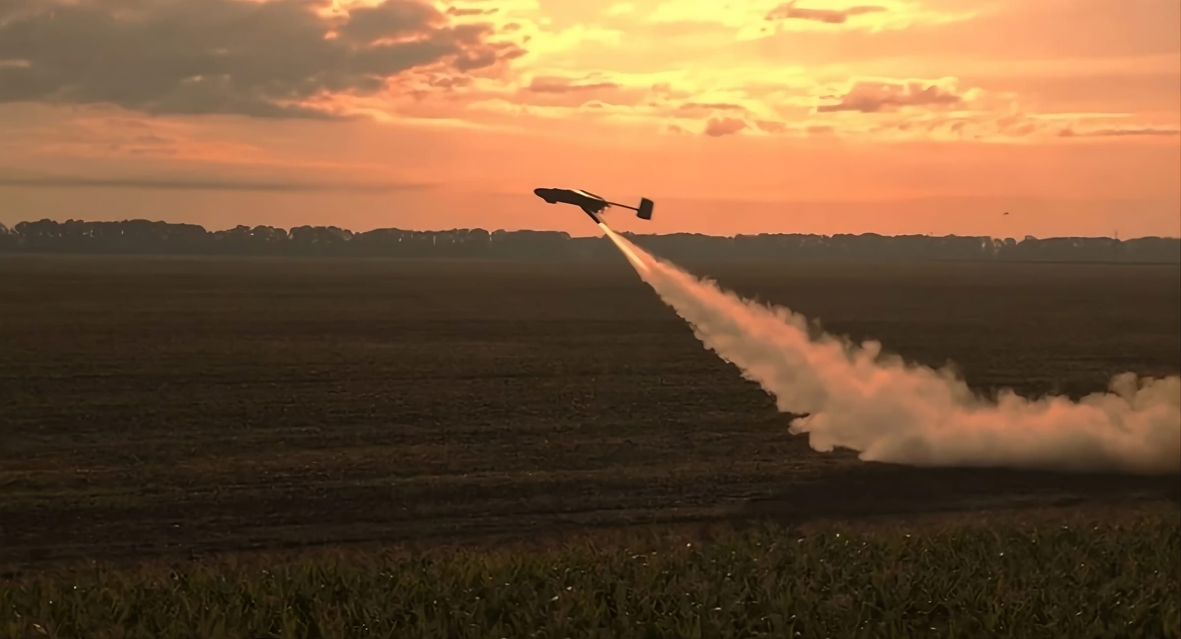 Dron de ala fija despegando desde el suelo impulsado por un cohete de aceleración, con estela de humo al atardecer.