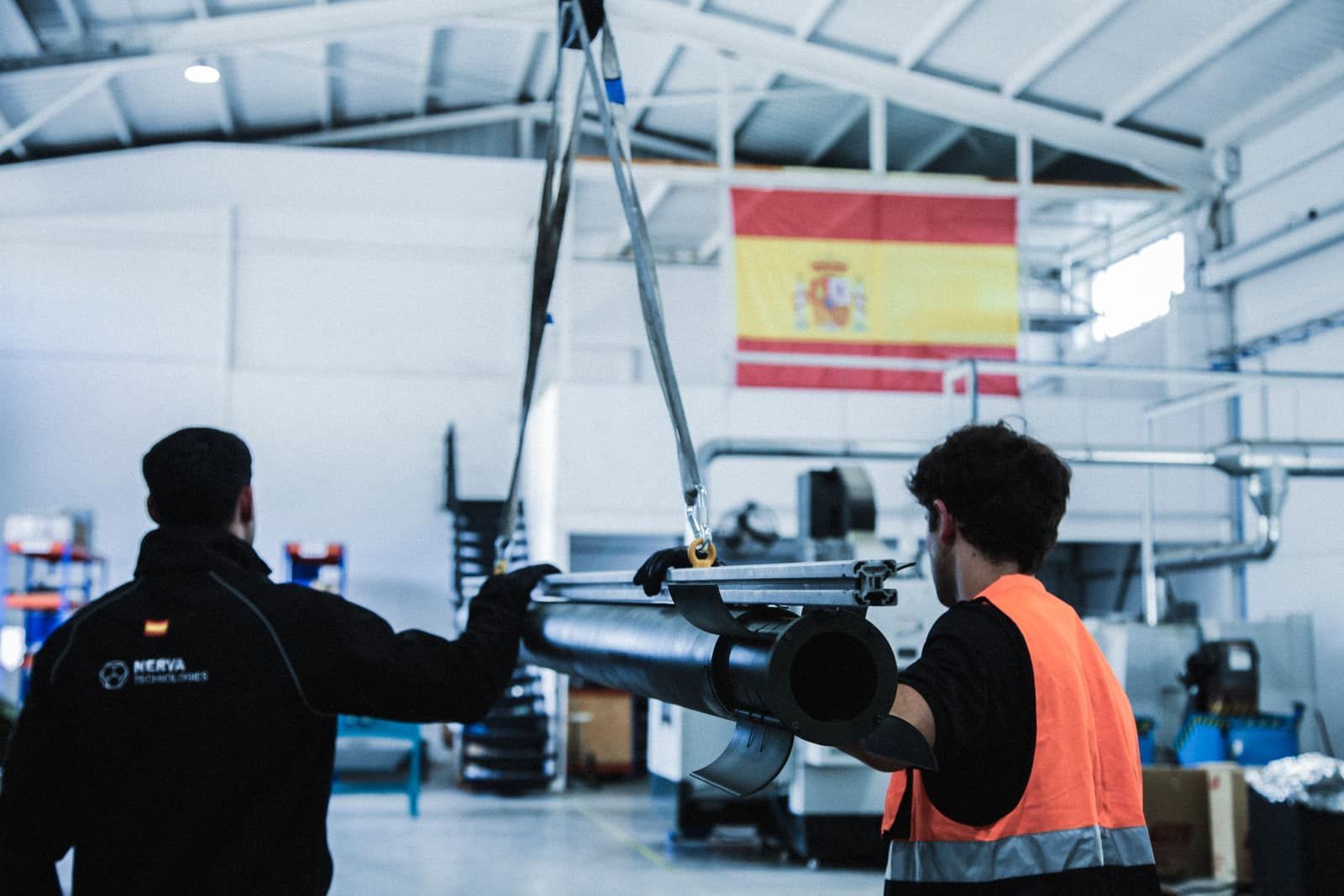 Dos operarios de Nerva guían un tubo de motor cohete suspendido de un puente grúa, con la bandera de España al fondo del taller.
